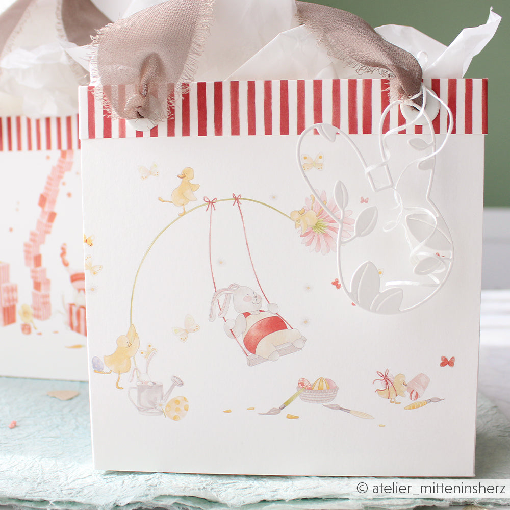 Geschenktüte von Alexandra Renke mit roten Streifen, Häschen auf einer Schaukel und pastellfarbenen Tiermotiven; mit einem durchsichtigen Bandhenkel versehen - ideal für kreative Papierprojekte oder passend zu Grußkarten.