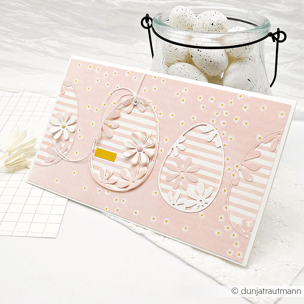 Eine rosafarbene Osterkarte mit gestreiften und floralen Eimotiven von Alexandra Renke Designpapier liegt neben einem Glas mit weiß gesprenkelten Eiern - die perfekte Inspiration für Papierprojekte.
