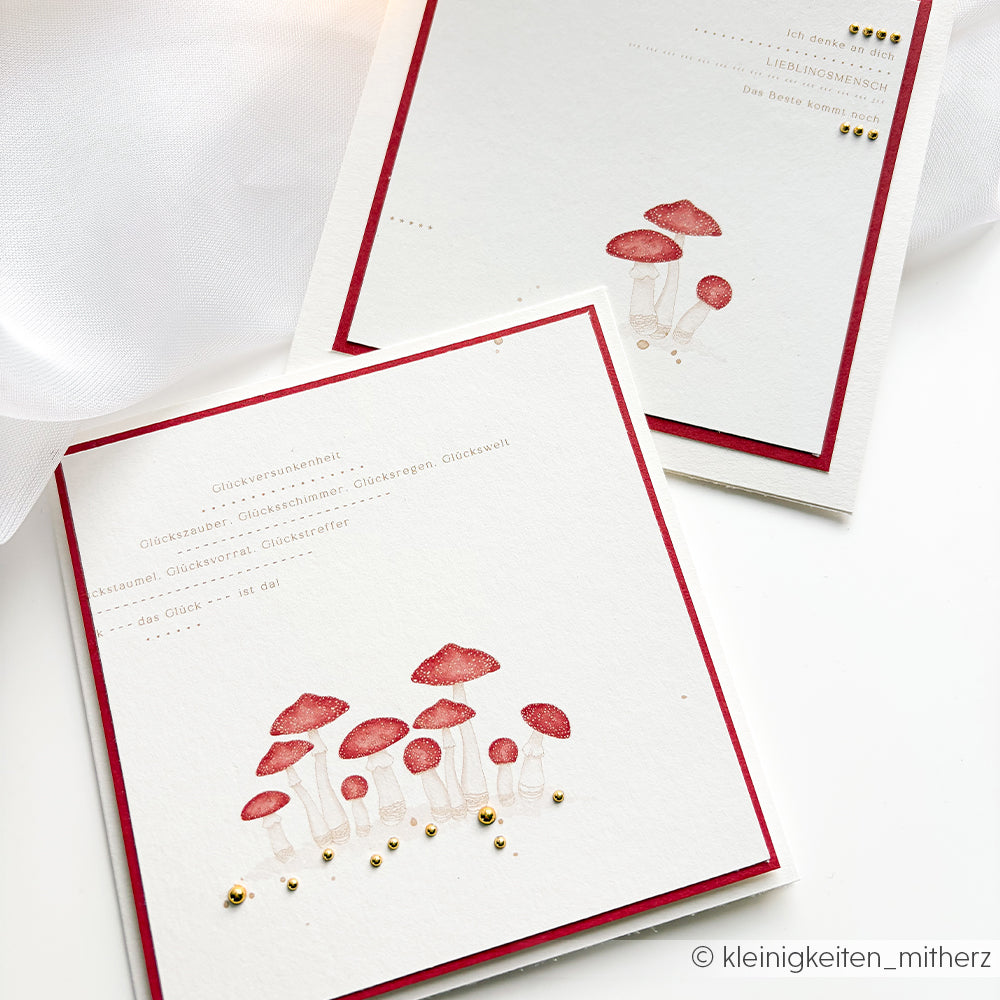 Zwei handgefertigte Grußkarten, verziert mit Pilzmotiven in Aquarelltechnik und goldenen Verzierungen auf strukturiertem Designpapier.