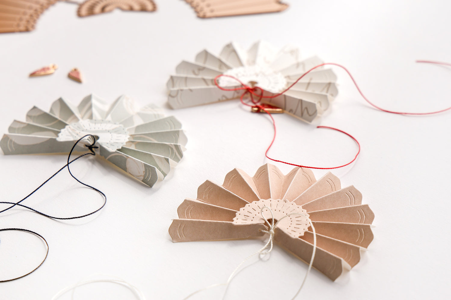 Drei kunstvoll gefaltete Papierfächer in verschiedenen Farben, die mit feinen Linien geprägt sind und an unterschiedlich farbigen Kordeln hängen. Die Fächer sind auf einem hellen Untergrund arrangiert und zeigen die Verwendung einer Stanzschablone für Bastelprojekte.