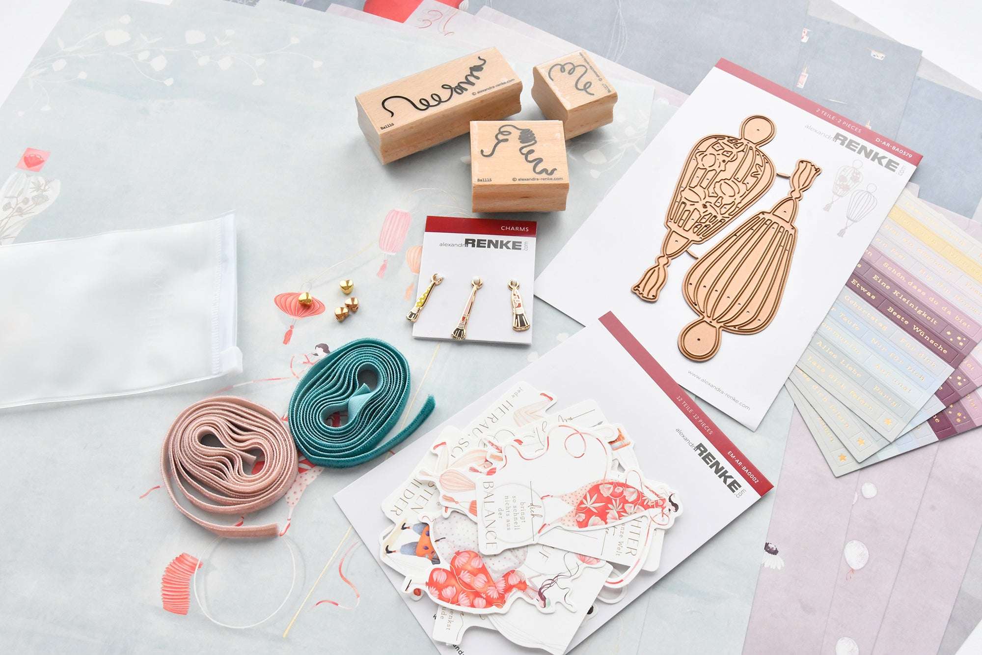 Verschiedenes Bastelmaterial wie Stempel, Metallstempel, Bänder, Anhänger und dekoratives Papier auf einer weißen Oberfläche.