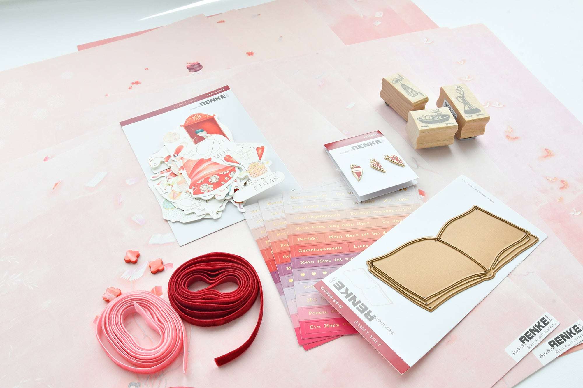 Verschiedenes rosa und rotes Bastelmaterial, Papier, Aufkleber, Stempel und Bänder auf einer hellen Fläche angeordnet.
