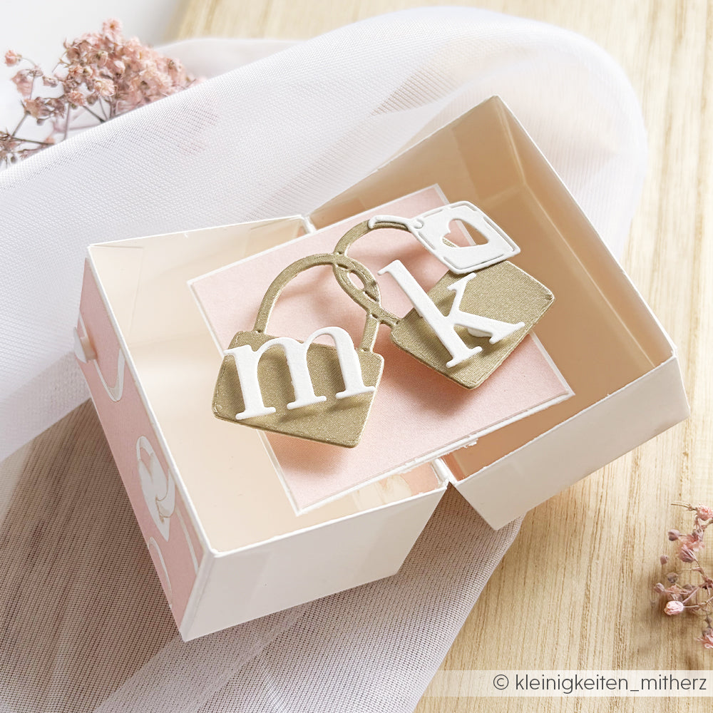 Geöffnete Geschenkbox mit zwei goldenen Schlüsselanhängern in Handtaschenform, jeweils mit weißem "m" und "k" - perfekt, um Alexandra Renke Motivstanze Schlosspaar zu verzaubern oder mit kreativen Designpapieren zu kombinieren.