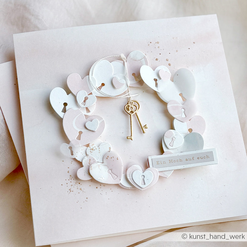 Eine handgefertigte Karte von Alexandra Renke zeigt einen herzförmigen Kranz mit Schlüssellochausschnitten, gefertigt aus Motivstanze Schlüssellochherzen und hochwertigen Designpapieren, sowie zwei goldene Schlüssel in der Mitte.