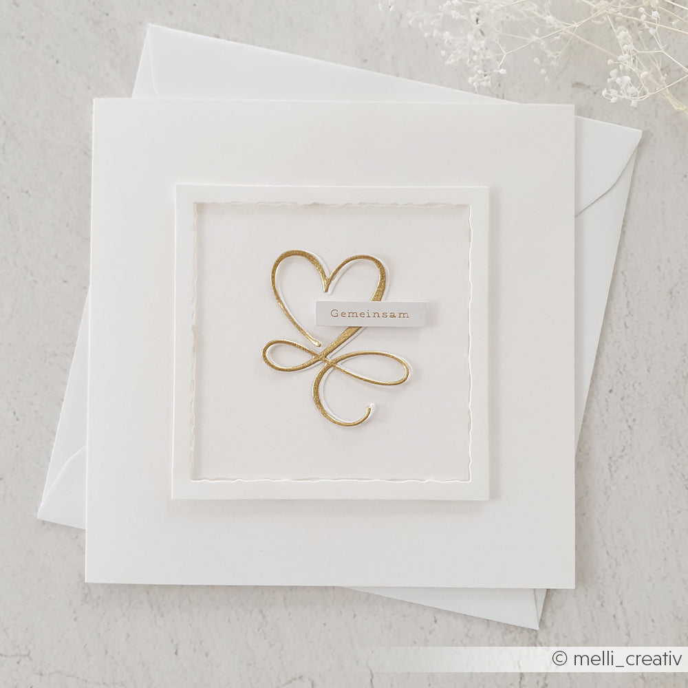 Weiße quadratische Karte von Alexandra Renke mit einem goldenen Herz und einer Schleife mit "Gemeinsam" auf strukturiertem Papier mit Motivstanze Quadrat, auf einem weißen Umschlag.