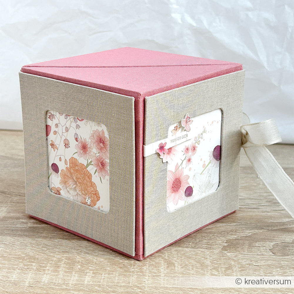 Dekorative rosa-beige Alexandra Renke PAPPKit Würfelbox mit Blumenmotiven, Fenstern und Schleifenband - ideal als Fotowürfelbox oder einzigartige Geschenkverpackung, die auf einer Holzfläche präsentiert wird.