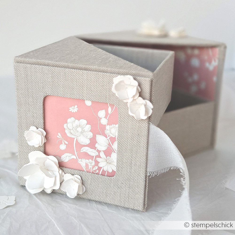 Eine quadratische graue Alexandra Renke PAPPKit-Geschenkbox mit einem rosafarbenen Blumenfenster, weißen Papierblumenverzierungen und einem Stoffband.