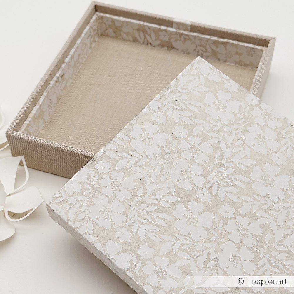Eine beige Einsteckbox von Alexandra Renke mit floralem, weißem Muster auf dem Deckel, teilweise geöffnet auf hellem Untergrund - ideal zum Selbermachen von Geschenkverpackungen.