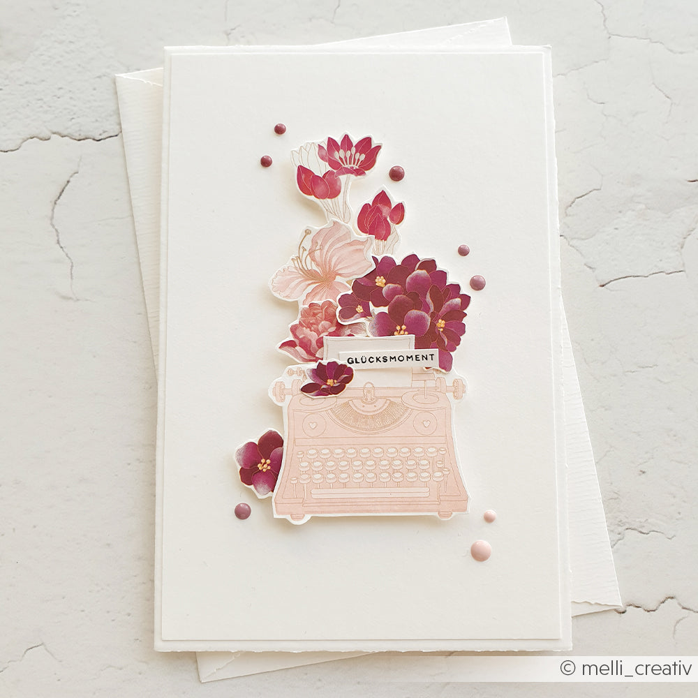 Eine Karte mit einer rosafarbenen Schreibmaschine und violetten Blumen, die mit Alexandra Renke Rub-On Rosarote Blütenpoesie für Bastelprojekte gestaltet wurde, zeigt das Wort GLÜCKSMOMENT auf weißem Strukturpapier.