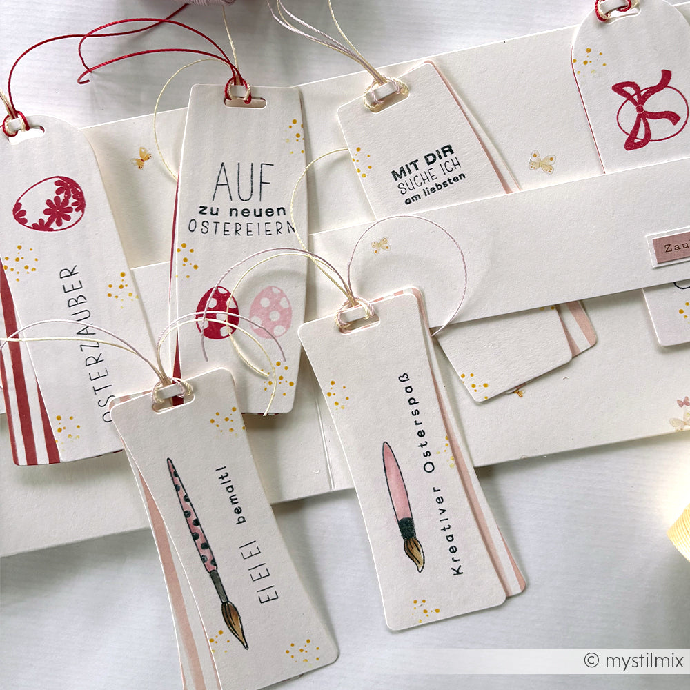 Handgefertigte Ostergeschenkanhänger von Alexandra Renke mit deutschen Grüßen und pastellfarbenen Illustrationen, hergestellt mit einer taillierten Etikett-Motivstanze, werden ordentlich auf einer weißen Fläche präsentiert.