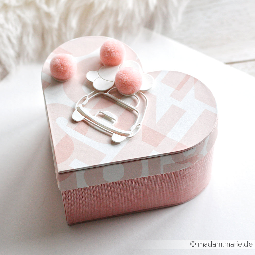 Rosafarbene Geschenkschachtel in Herzform von Alexandra Renke mit Bommeln, einer weißen Blume und einem Geldbeutel auf dem Deckel, hergestellt aus Designpapieren mit eleganten DieCut-System-Details, ausgestellt auf einer hellen Oberfläche.