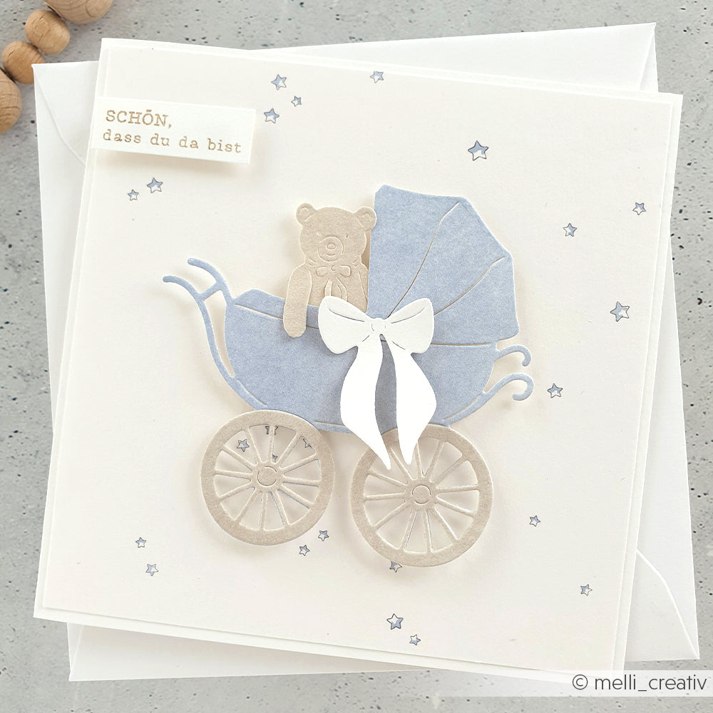 Handgefertigte Karte aus Alexandra Renke-Papier mit einem blauen Kinderwagen, der mit einer Kinderwagenstanze gestanzt wurde, sowie einem Teddybär, einer weißen Schleife und einem kleinen Stern als Dekoration.