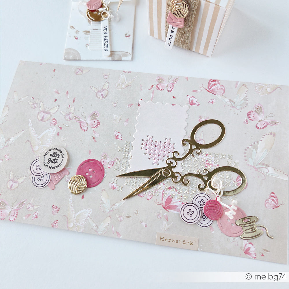 Flatlay mit Zierscheren, Knöpfen, rosa Bastelartikeln sowie dem „Spaß mit Wolle“-Anhänger und der Motivstanze von Alexandra Renke auf einem Hintergrund mit Schmetterlingsmuster.