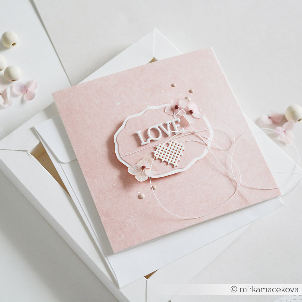 Rosa handgefertigte Karte mit der Aufschrift „LOVE“, kleinen Blumen und einer Schnur, auf einem weißen Umschlag angebracht. Hergestellt aus eleganten Designpapieren von Alexandra Renke und veredelt mit dem Stanzsystem „Stickrahmen Oval“ von Alexandra Renke für ein bezauberndes, detailreiches Finish.