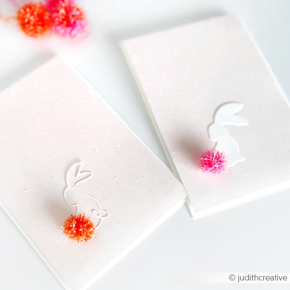 Zwei weiße Karten mit Hasensilhouetten und bunten Bommelschwänzen, hergestellt mit Rico Design robustes Garn für eine strukturierte Oberfläche, auf weißem Hintergrund.