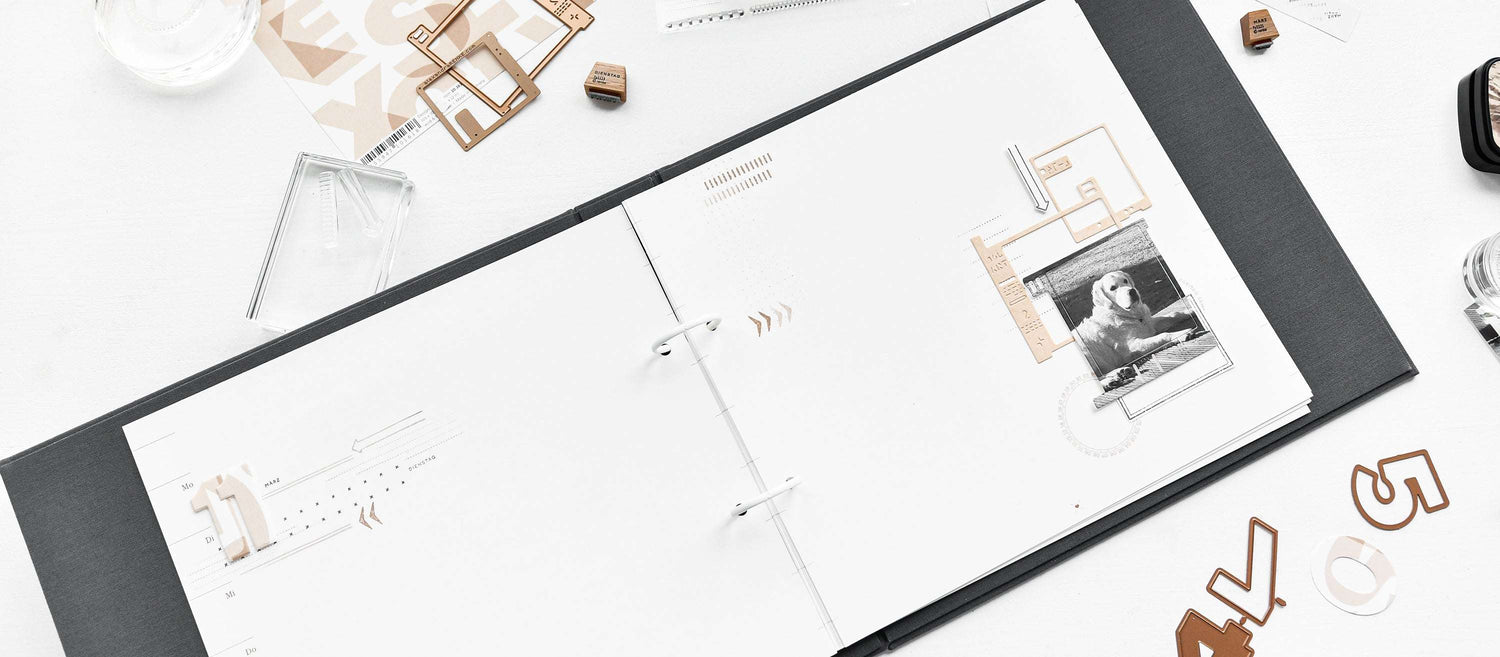 Offene Mappe mit minimalistischen Scrapbook-Seiten, Bastelmaterial und einem Foto auf einem weißen Schreibtisch.