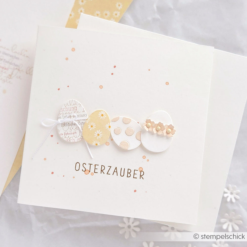 Quadratische Karte von Alexandra Renke mit einem Vier-Eier-Set-Motiv und dem Text OSTERZAUBER in der Mitte, akzentuiert mit kleinen orangefarbenen Punkten - eine reizvolle Ergänzung für Ihre Designpapiere-Projekte.