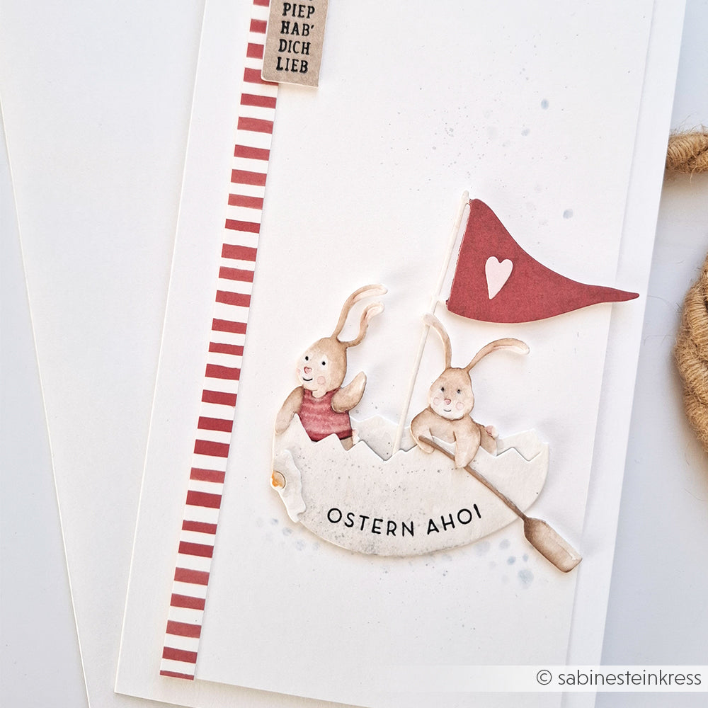 Zwei Cartoon-Hasen in einem Eierschalenboot mit roter Flagge und dem Text "OSTERN AHOI" auf einer gestreiften weißen Karte von Alexandra Renke - ideal für DieCut Systeme oder die Motivstanze Ostern, um einzigartige Osterbasteleien zu gestalten.