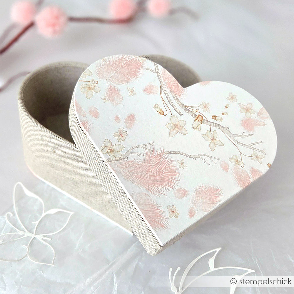 Alexandra Renke herzförmige Geschenkverpackung mit zartrosa Blumen- und Federmotiv, teilweise geöffnet auf einem eleganten hellen Hintergrund.