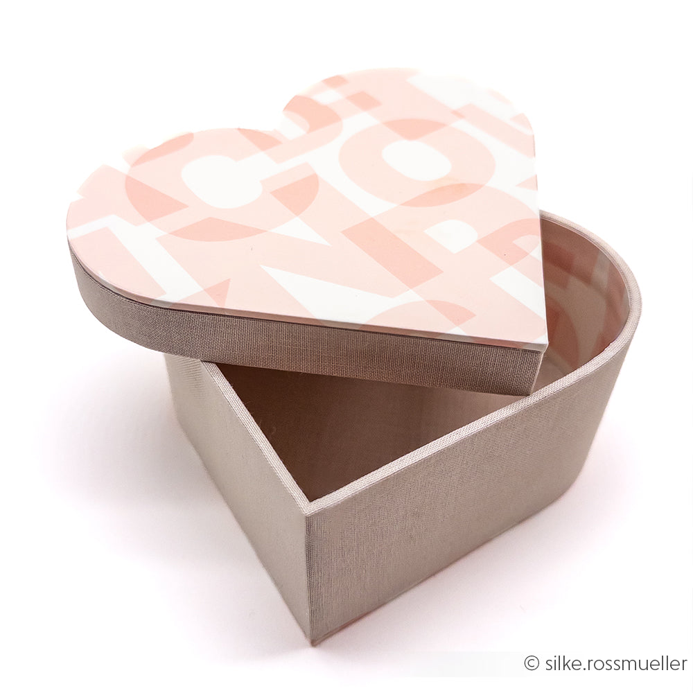 Eine herzförmige Box von Alexandra Renke mit rosa-weiß gemustertem Deckel, teilweise geöffnet auf weißem Hintergrund - ideal für Geschenkverpackungen oder als Inspiration für Ihr nächstes Herzbox basteln Projekt.