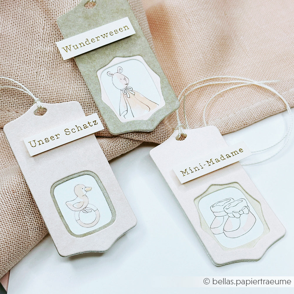 Drei pastellfarbene Geschenkanhänger von Alexandra Renke mit deutschem Text und niedlichen Baby-Illustrationen, ideal für Bastelprojekte oder Spiel und Kuschelspaß, werden auf einer beigen Stoffoberfläche präsentiert.