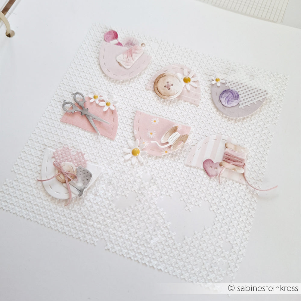 Eine Scrapbook-Seite mit pastellfarbenen Verzierungen zum Thema Nähen – Garn, Schere, Knöpfe, Faden, Gänseblümchen – und einem Kreuzstich-Hintergrund, der mit wiederverwendbaren Schablonen von Alexandra Renke gestaltet wurde und für kreative Akzente sorgt.
