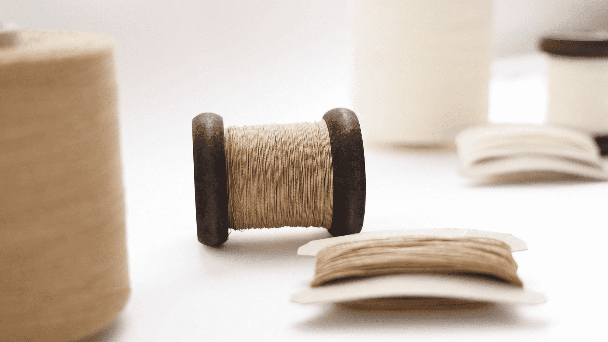 Eine Holzspule mit beigem Garn, umgeben von anderen Spulen und Garnkarten auf einer weißen Fläche.