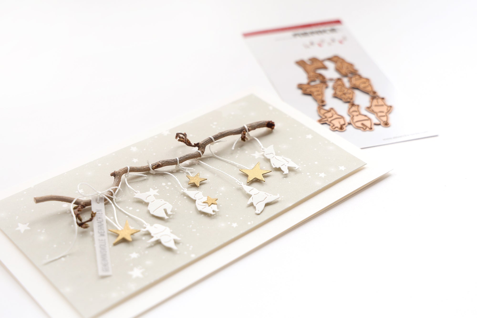 Handgemachte Karte mit hängenden weißen Tieren und goldenen Sternen an einem Zweig, mit Bastelmaterial in der Nähe.