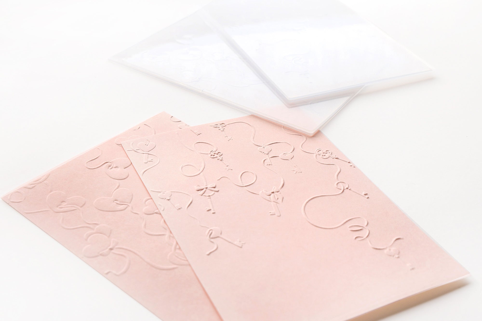 Zwei hellrosa geprägte Karten mit Blumenmotiven, daneben zwei transparente Umschläge auf einer weißen Fläche.