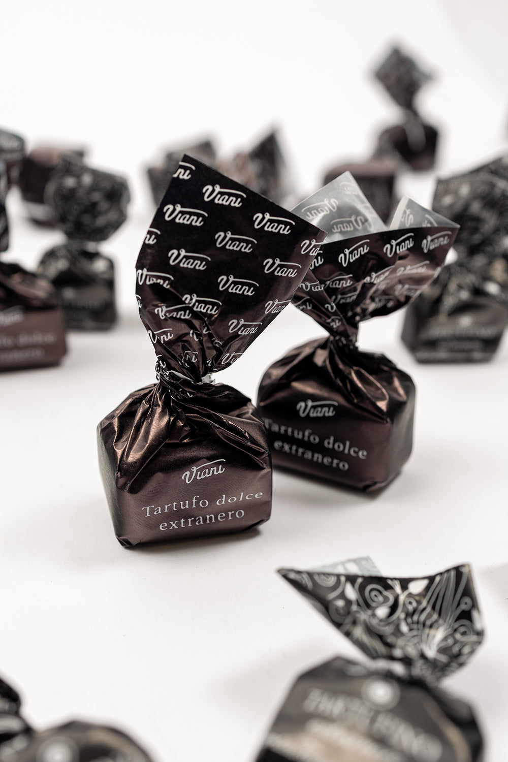 Zwei Viani Tartuffo Dolce Extranero-Pralinen in braunen Markenverpackungen, umgeben von weiteren vereinzelten, verpackten Pralinen.