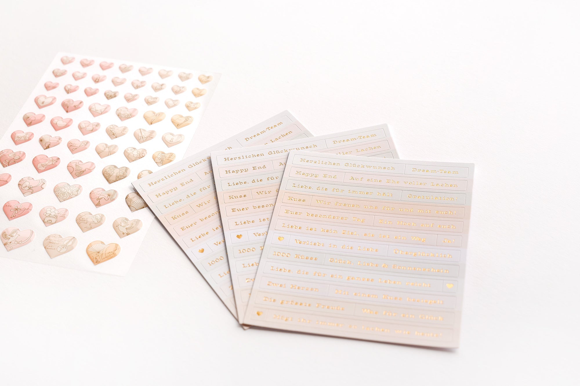Fünf Bögen mit dekorativen Aufklebern, vier mit goldenem Text und einer mit verschiedenen Herzmotiven auf einer weißen Oberfläche.