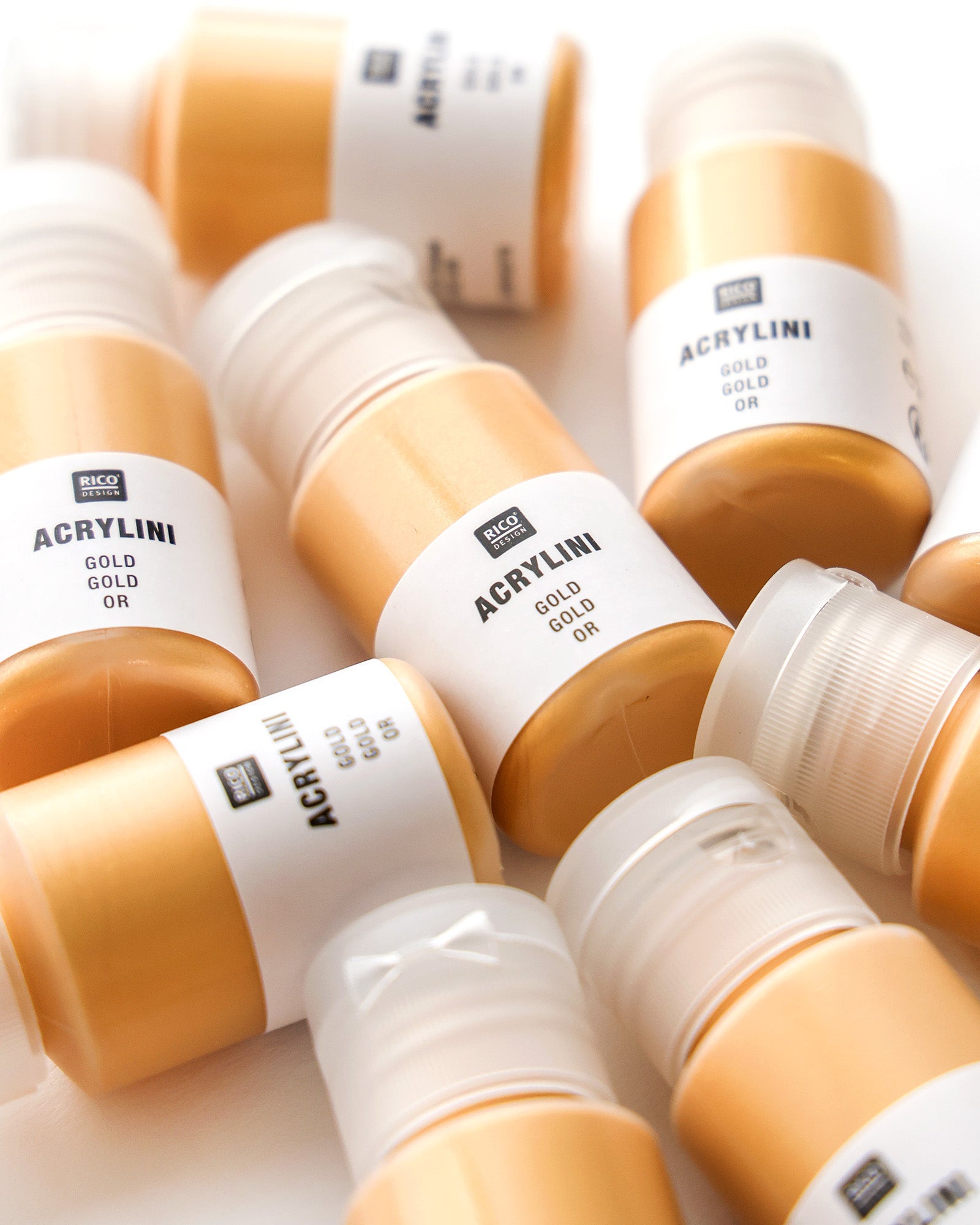 Mehrere Flaschen goldener Acrylfarbe mit der Aufschrift ACRYLINI GOLD sind auf einer weißen Fläche verstreut.