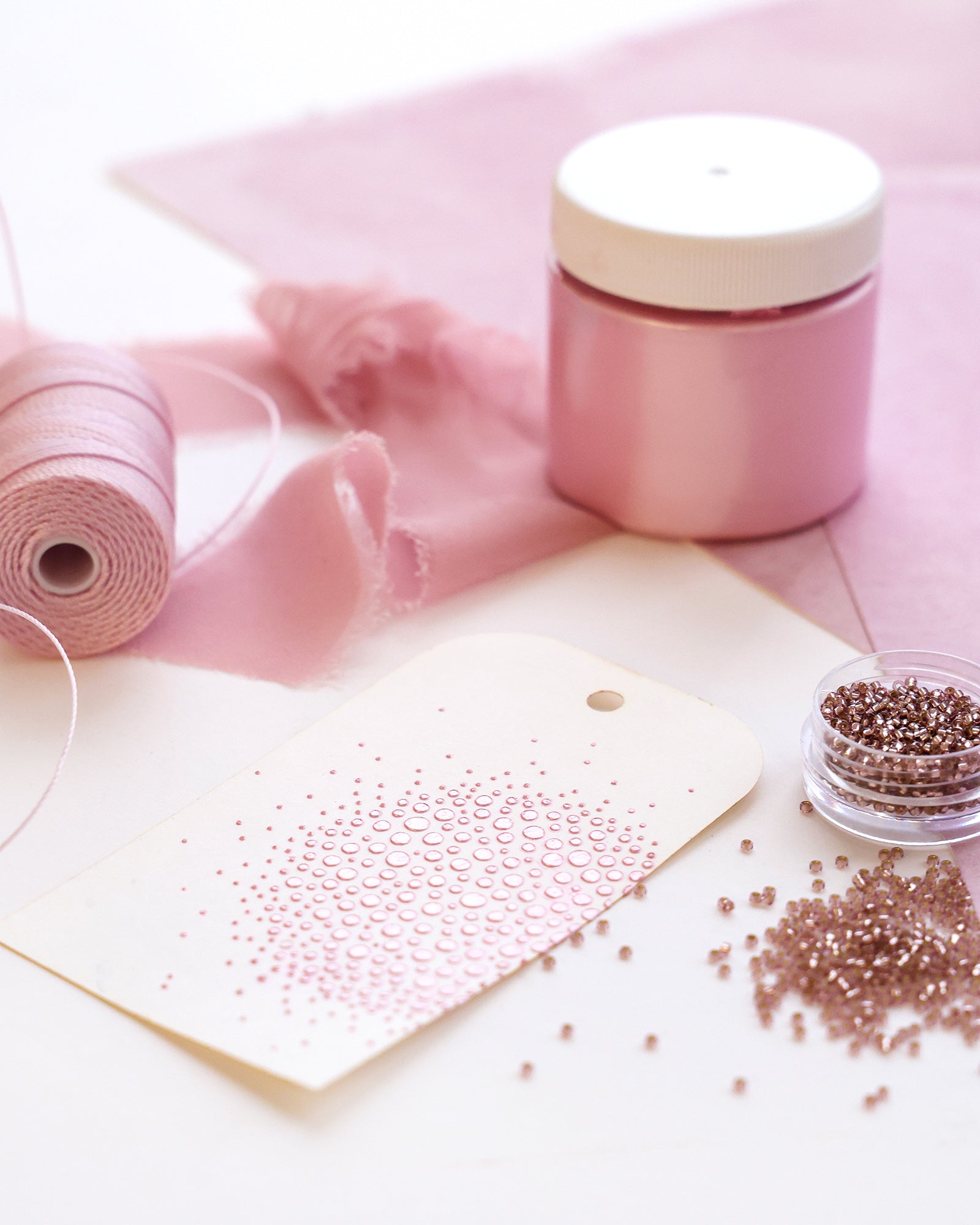 Rosa Bastelmaterialien, Perlen, Stoff und Schnur, die auf einem Tisch mit einem verzierten Anhänger im Vordergrund angeordnet sind.