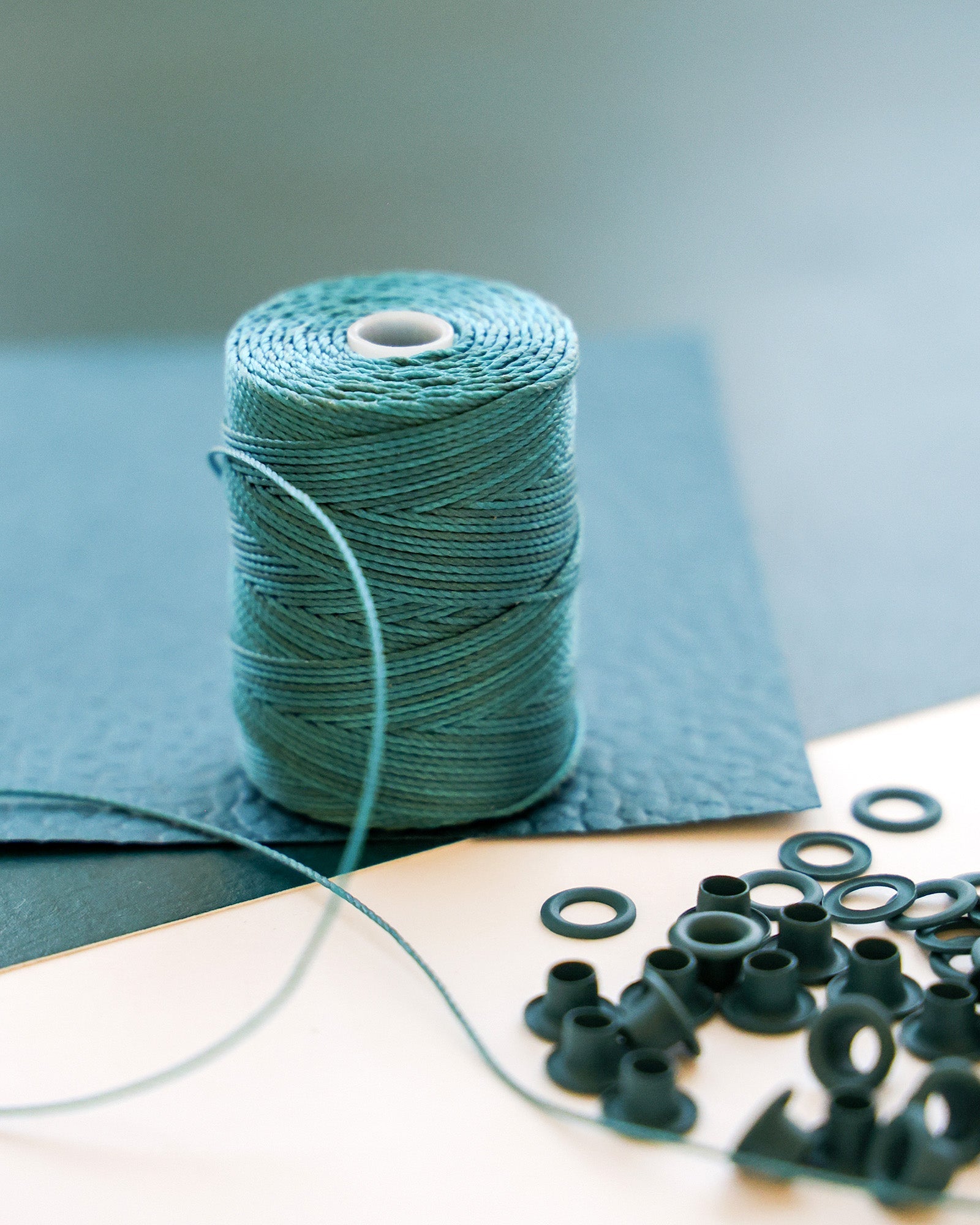 Eine Spule mit blaugrünem Garn mit kleinen schwarzen Ösen und blauem, strukturiertem Material im Hintergrund.