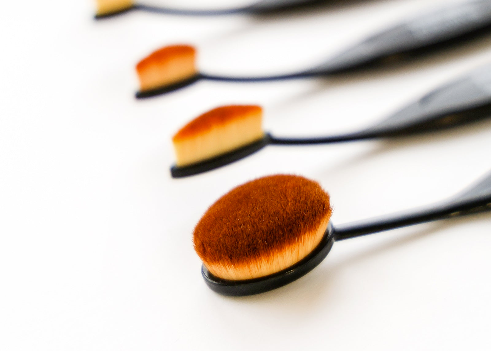 Sechs ovale Schminkpinsel mit schwarzen Griffen, die in einer diagonalen Linie auf weißem Hintergrund angeordnet sind.