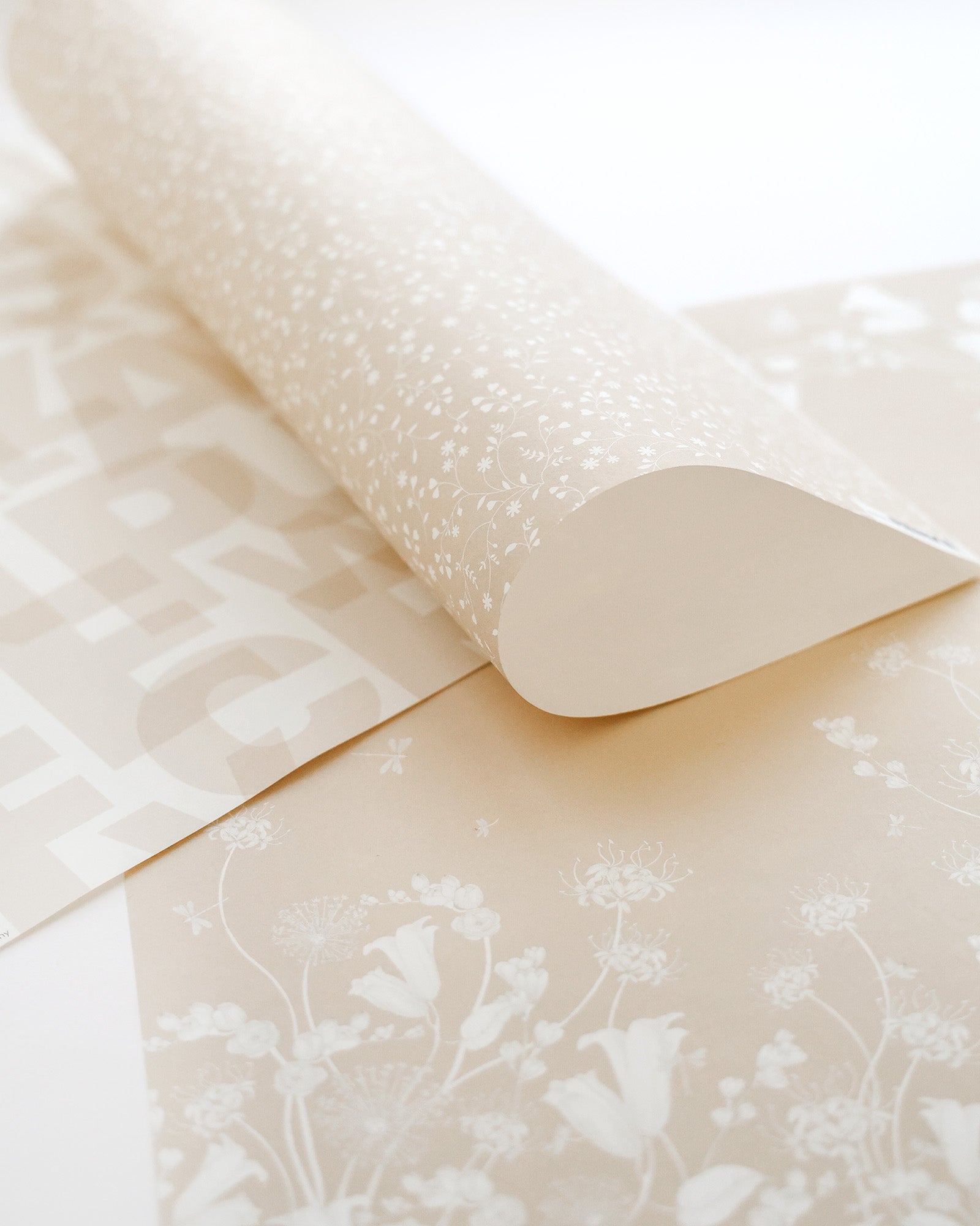 Nahaufnahme von beige gemusterten Tapetenbahnen mit Blumen- und Buchstabenmustern, eine Bahn teilweise aufgerollt.