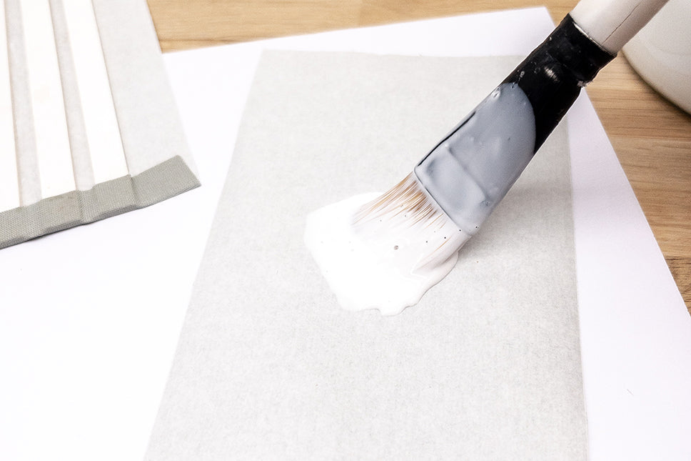 Ein Pinsel, der weißen Leim auf ein weißes Blatt Papier auf einem Holztisch aufträgt.