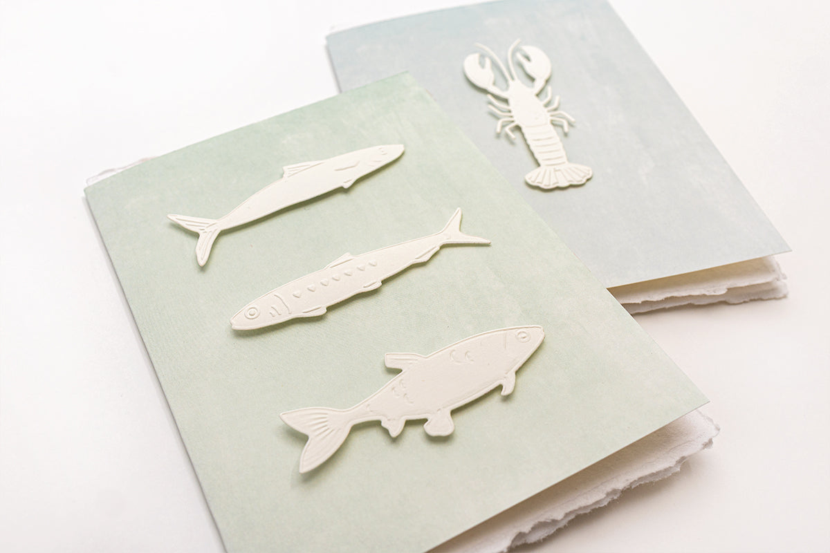Blätter aus Dekorationspapier mit pastellfarbenen Meeresmotiven, darunter Fische, Muscheln und Segelboote, auf weißem Hintergrund.