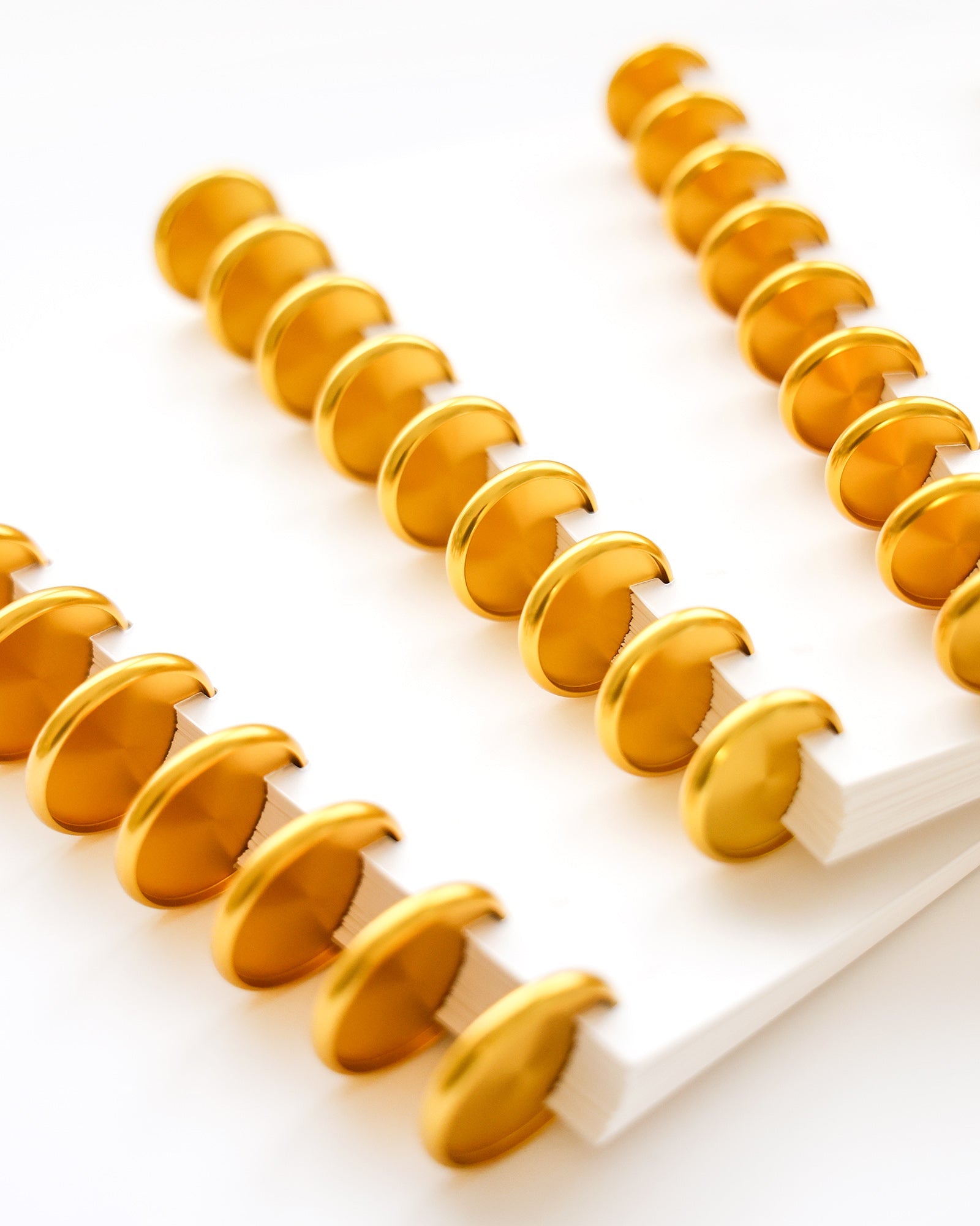 Nahaufnahme von weißen Notizbüchern mit goldglänzenden Einbandringen, die diagonal auf einer weißen Fläche angeordnet sind.