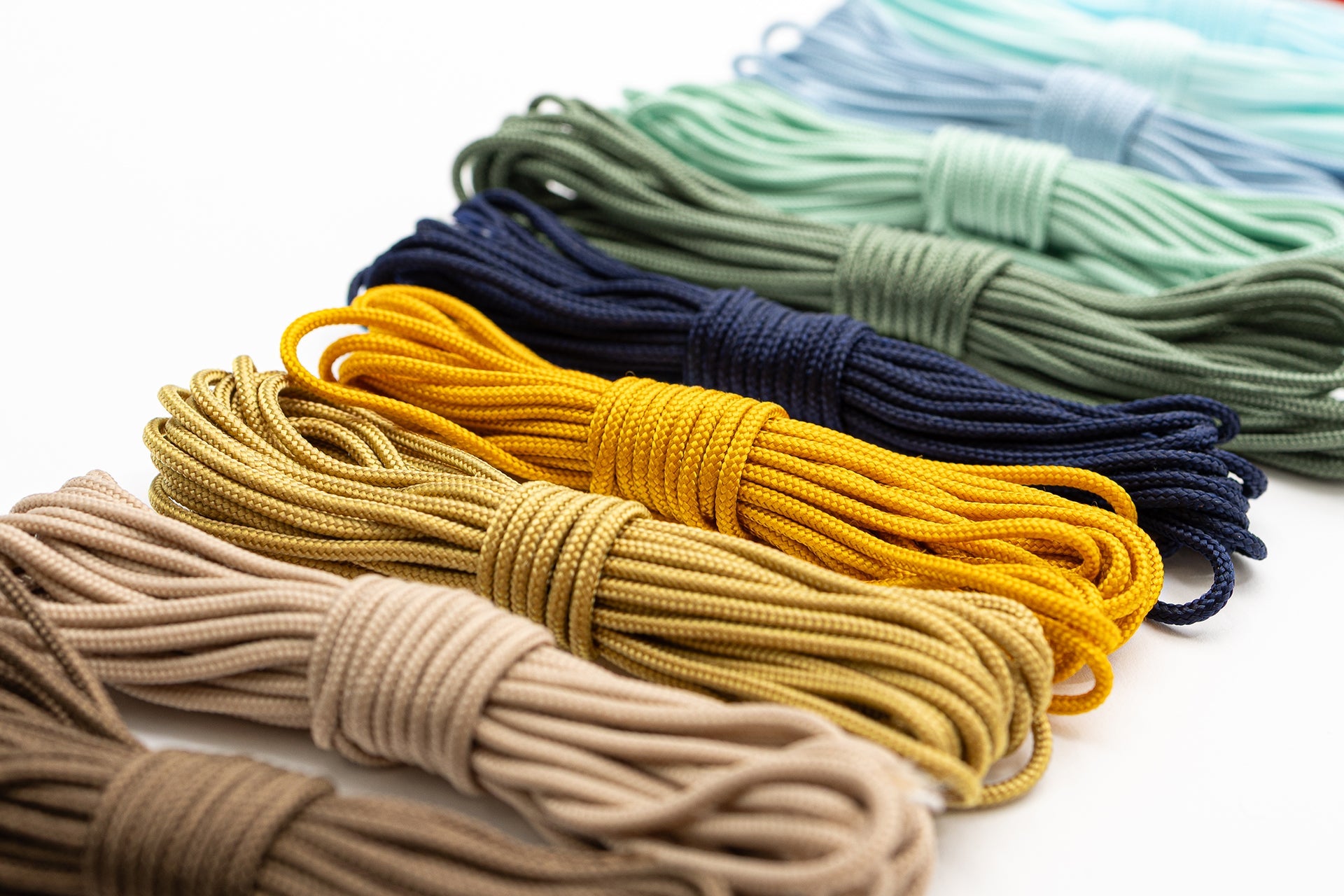 Paracord-Bündel in Beige, Gelb, Marineblau, Grün, Hellblau und Türkis sind ordentlich in Reihen auf einer weißen Fläche angeordnet.