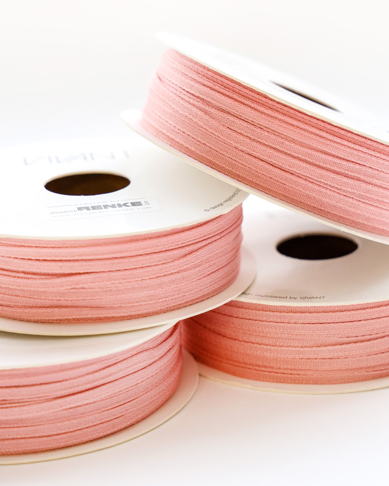 Vier Spulen mit zartem rosa Band, gestapelt auf einer weißen Fläche, wobei zwei Spulen auf der Seite liegen.