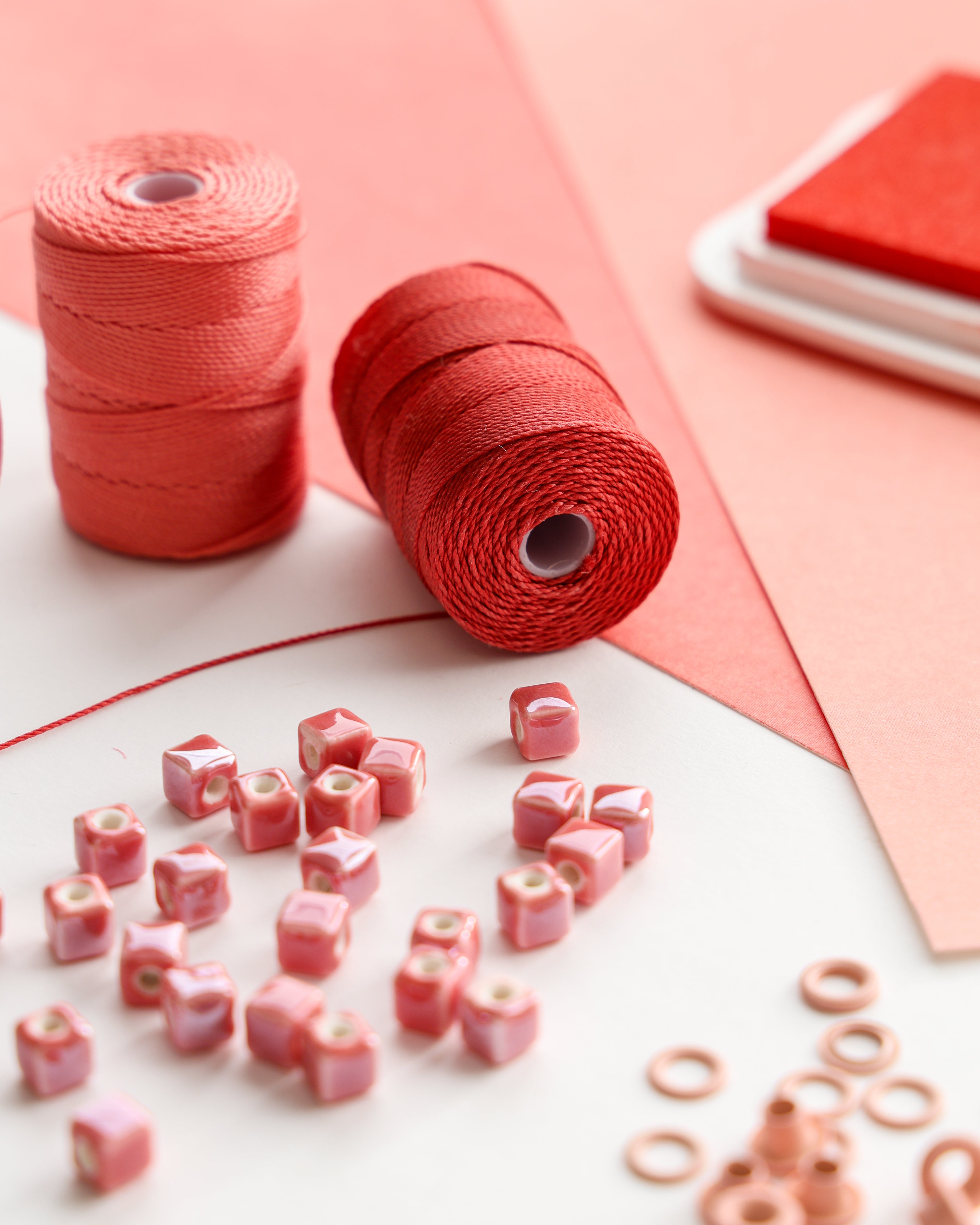 Korallenfarbene Fadenspulen, rosafarbene quadratische Perlen und Papierbögen, die auf einer weißen und rosafarbenen Fläche angeordnet sind.