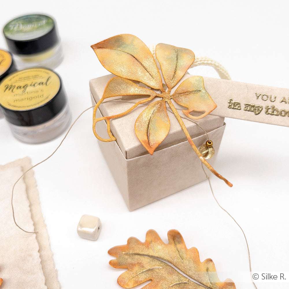 Geschenkschachtel mit Blattdekoration aus Metall, herbstlichem Etikett und Kunstzubehör in der Nähe auf einer weißen Oberfläche.