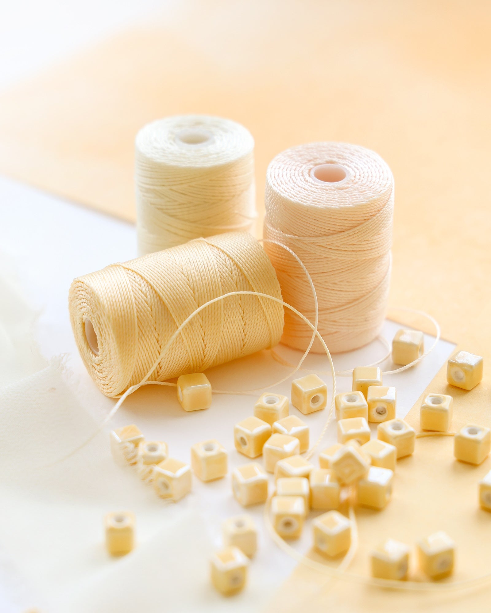 Drei Spulen mit cremefarbenem Garn und cremefarbenen quadratischen Perlen auf einer hellen Oberfläche mit einem weichen Hintergrund.