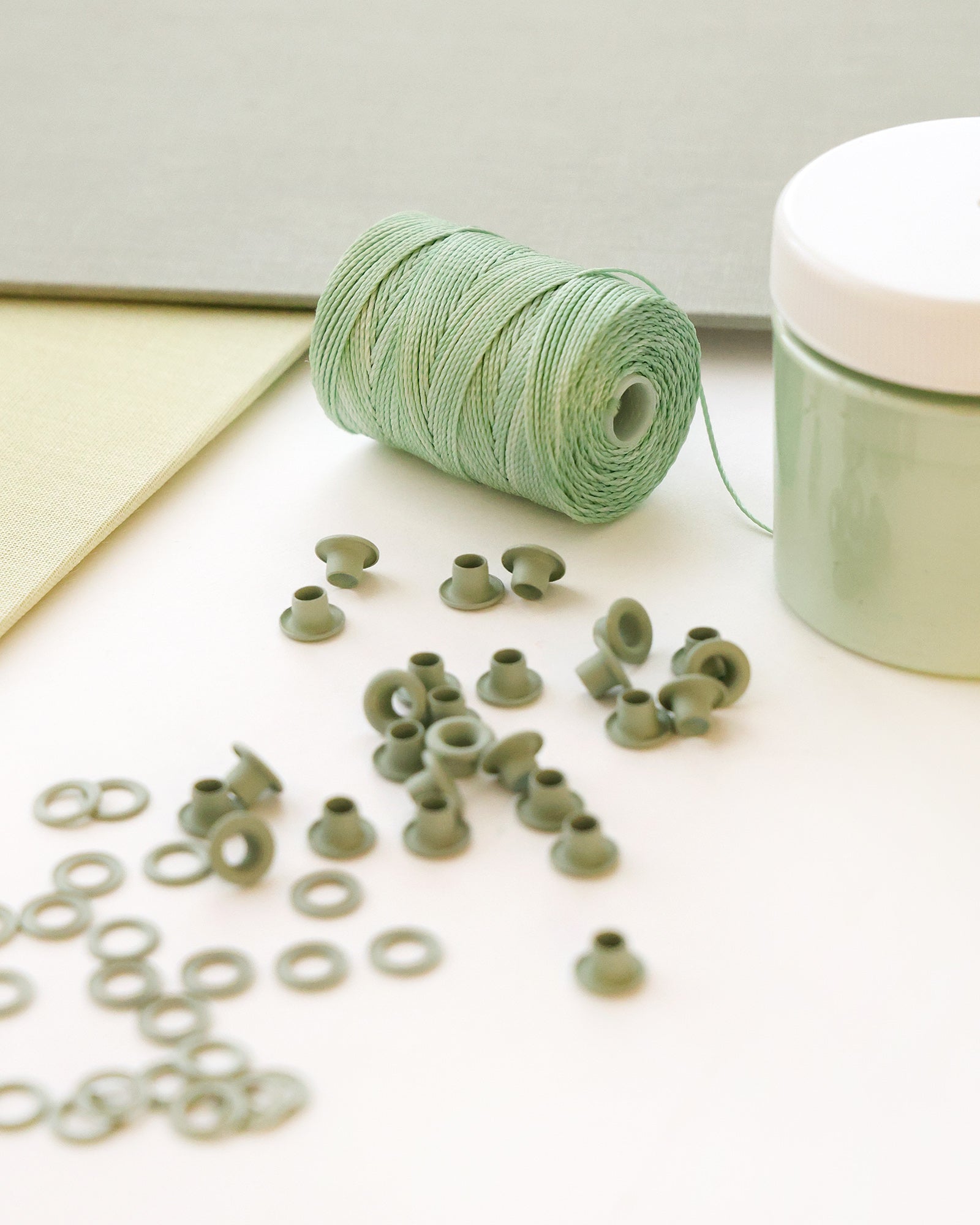 Eine Spule mit grünem Garn, kleine grüne Tüllen und ein Farbbehälter auf einer hellen Fläche.