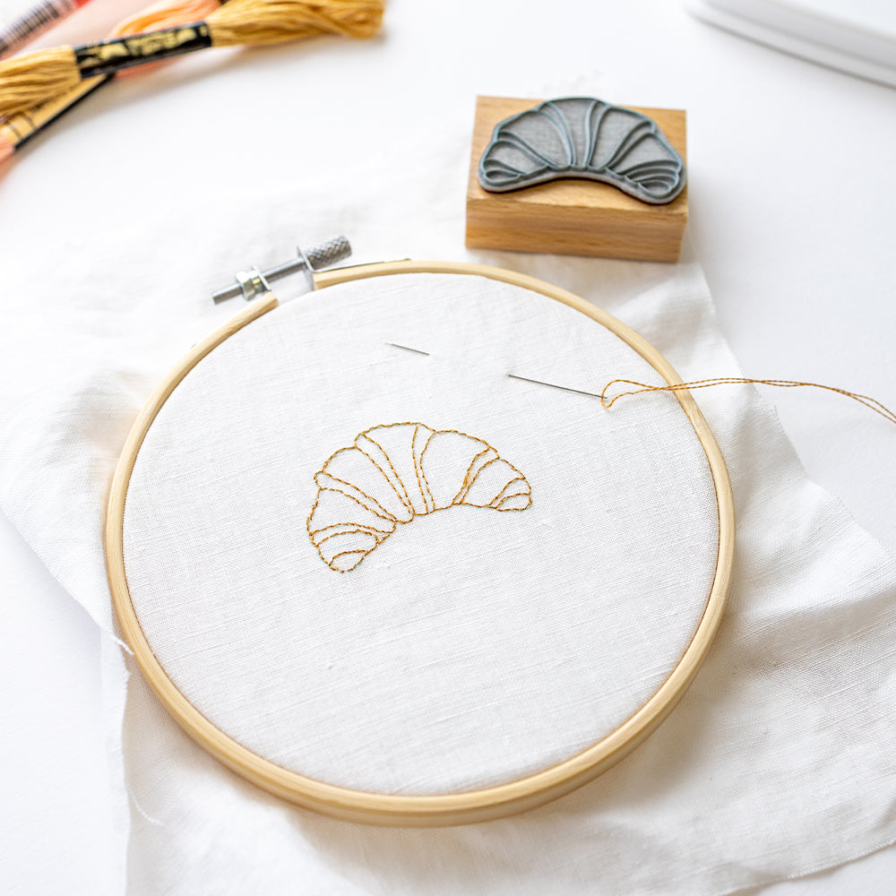 Stickrahmen mit dem Umriss eines gestickten Croissants, daneben Stempel und Garn.