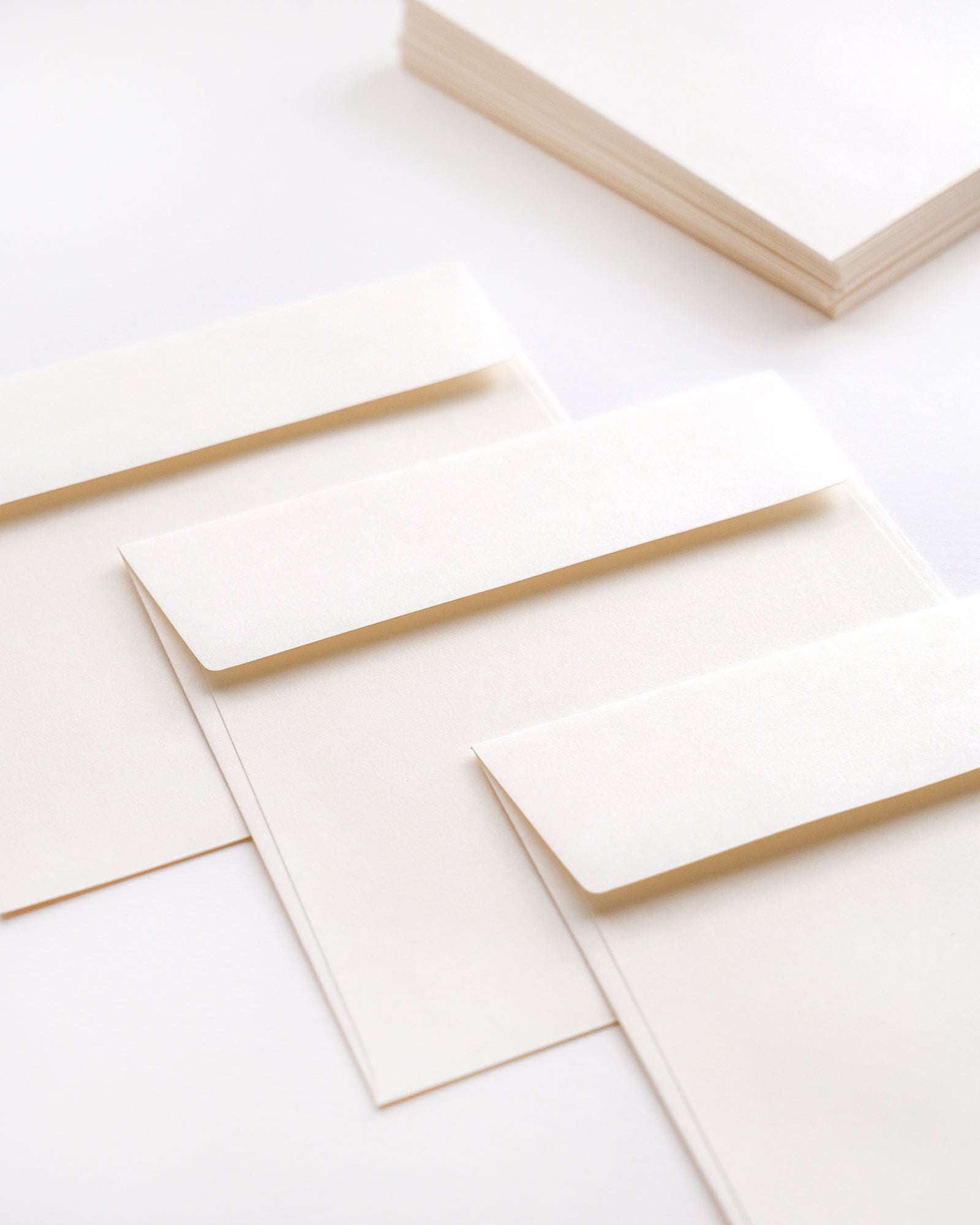 Drei cremefarbene Umschläge auf einer weißen Fläche, mit einem Stapel passenden Papiers im Hintergrund.