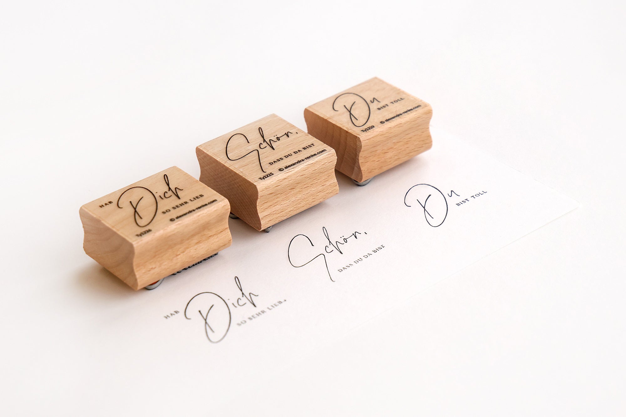 Drei Holzstempel mit elegantem handgeschriebenem Text, die neben gestempelten Abdrücken auf weißem Papier abgebildet sind.