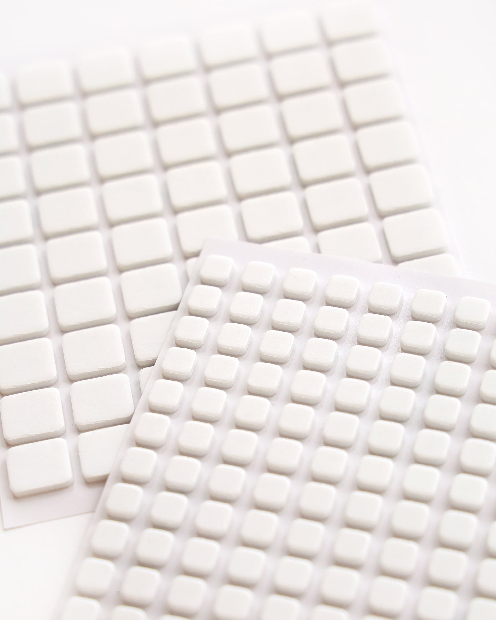 Nahaufnahme von weißen, quadratischen Klebepads aus Schaumstoff, die in Reihen auf Blättern angeordnet sind.