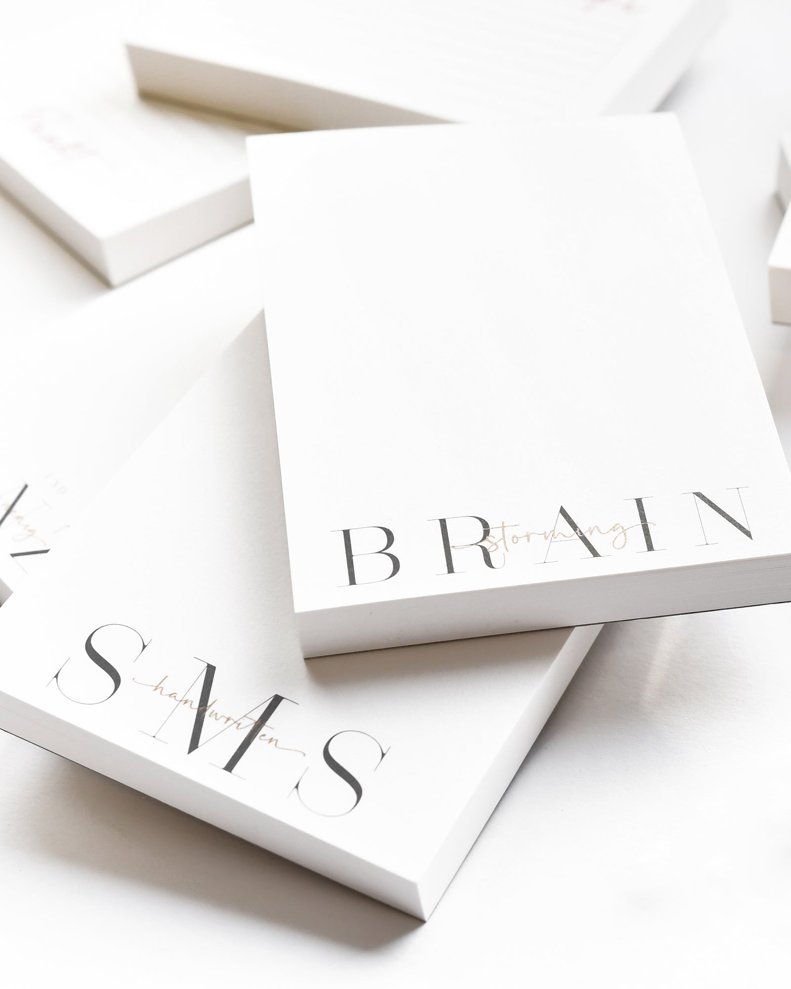 Weiße Notizblöcke, gestapelt, mit "BRAIN" und "SMS" in großen, eleganten Buchstaben am unteren Rand.